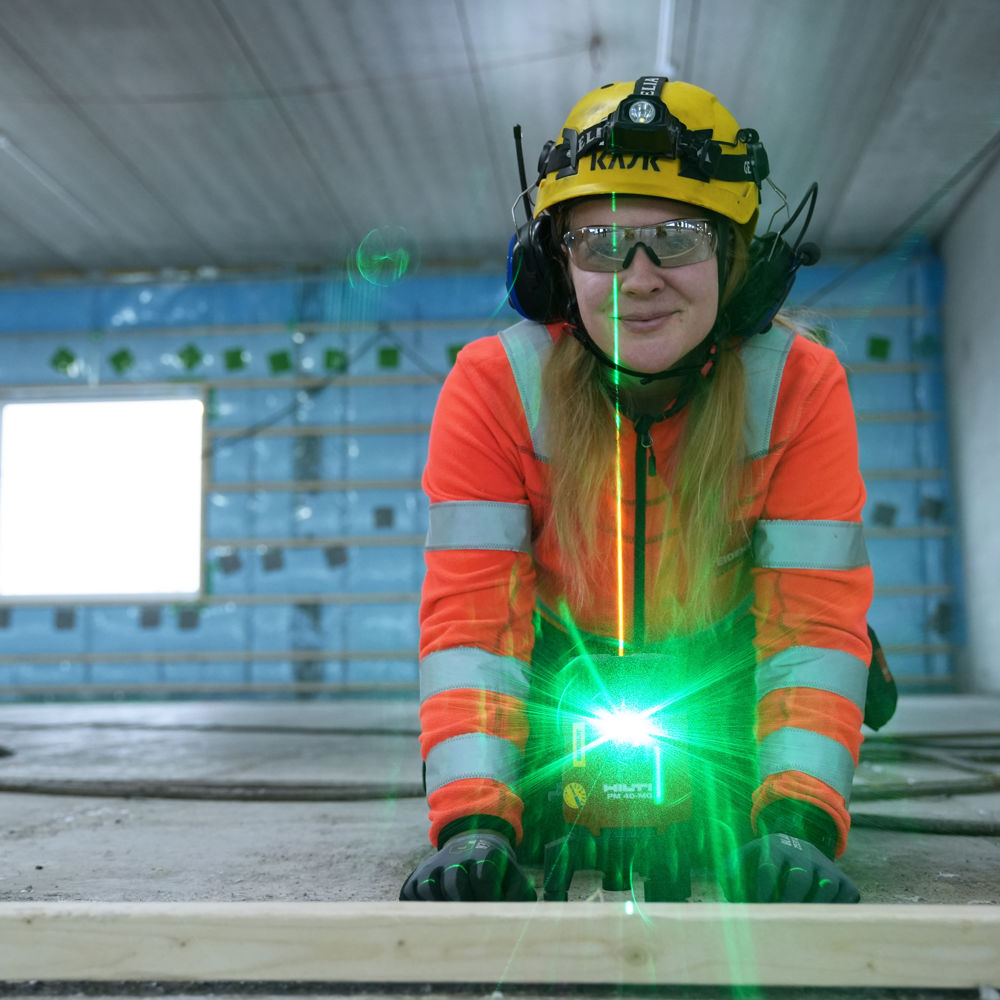 Ung kvinnlig byggarbetare i skyddsutrustning, Stella, står vid grönlysande laserpekare inomhus i ett nybuggnadsprojekt.