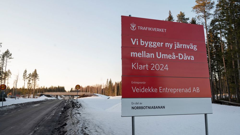 Snöklädd landsväg med viadukt. Förgrunden syns skylt med texten: "Vi bygger ny järnväg mellan Umeå-Dåva. Klart 2024. Trafikverket."