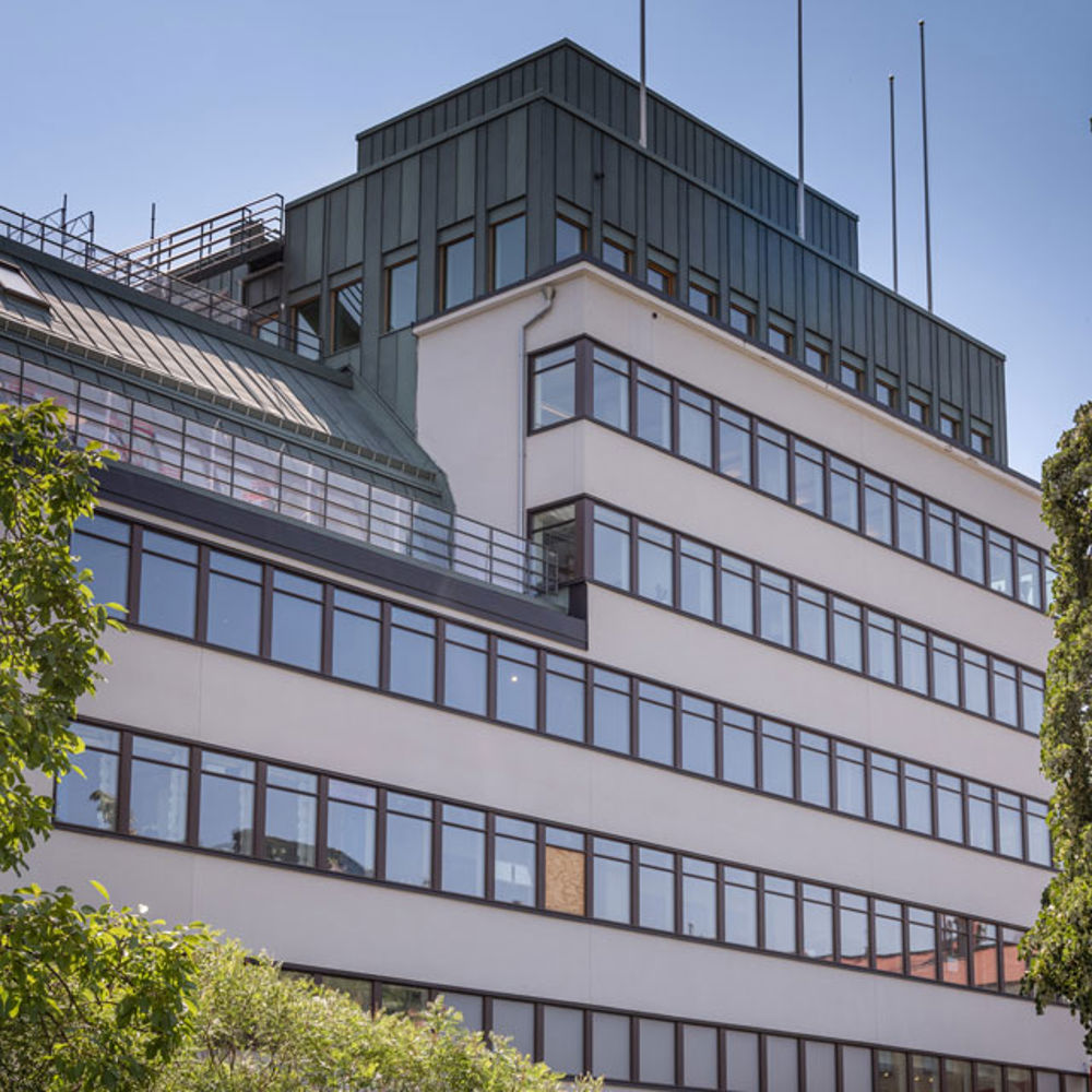 Ljus flervåningsbyggnad i funkisstil omgivet av gröna träd.