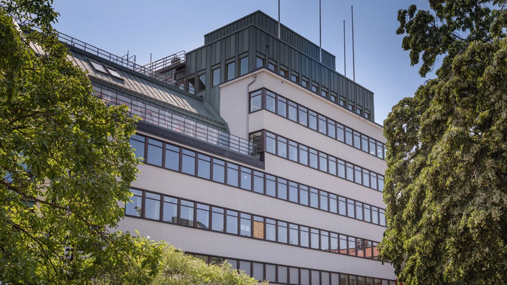 Ljus flervåningsbyggnad i funkisstil omgivet av gröna träd.