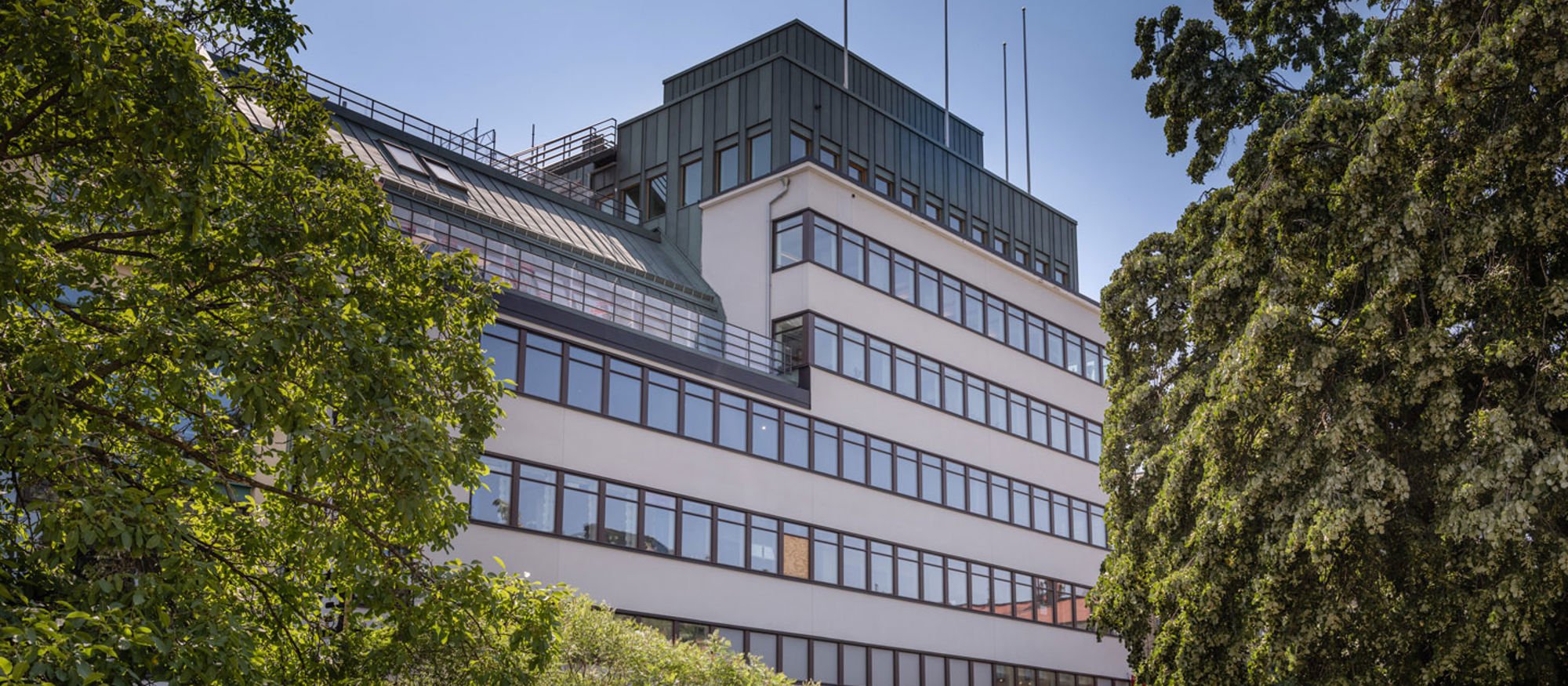 Ljus flervåningsbyggnad i funkisstil omgivet av gröna träd.