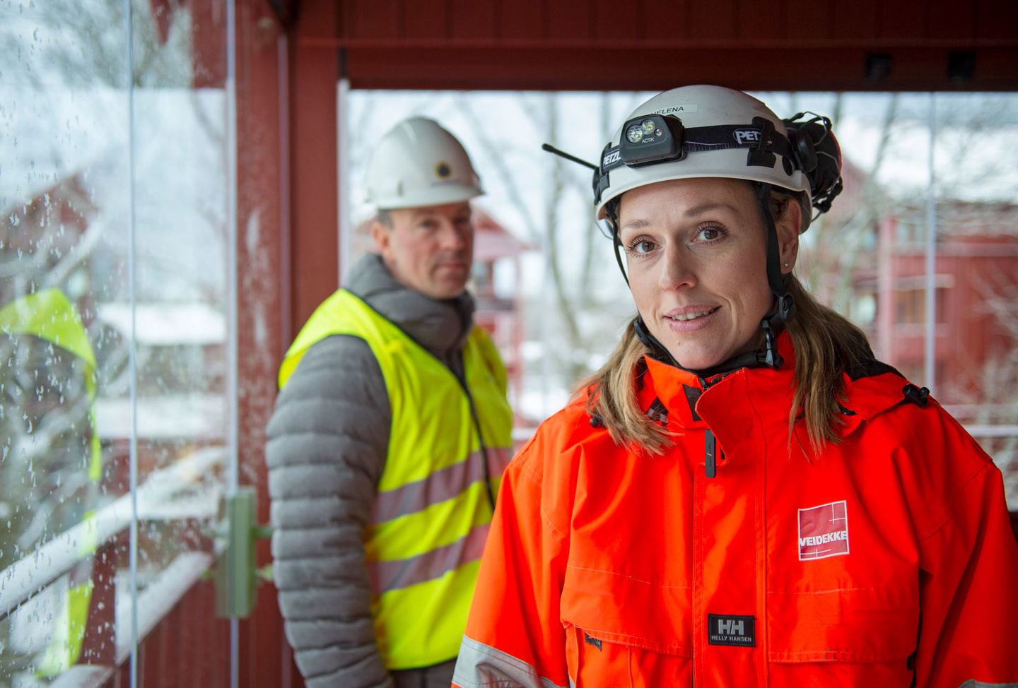 En kvinna står i förgrunden i varselkläder och bygghjälm, i bakgrunden står en man.