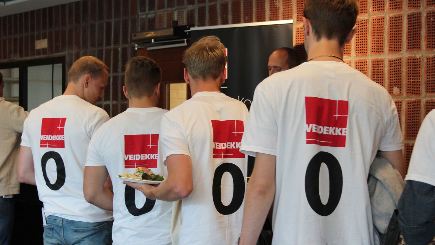 Fyra killar står i en kö med vita t-shirts med Veidekke-logon och en nolla på ryggen.