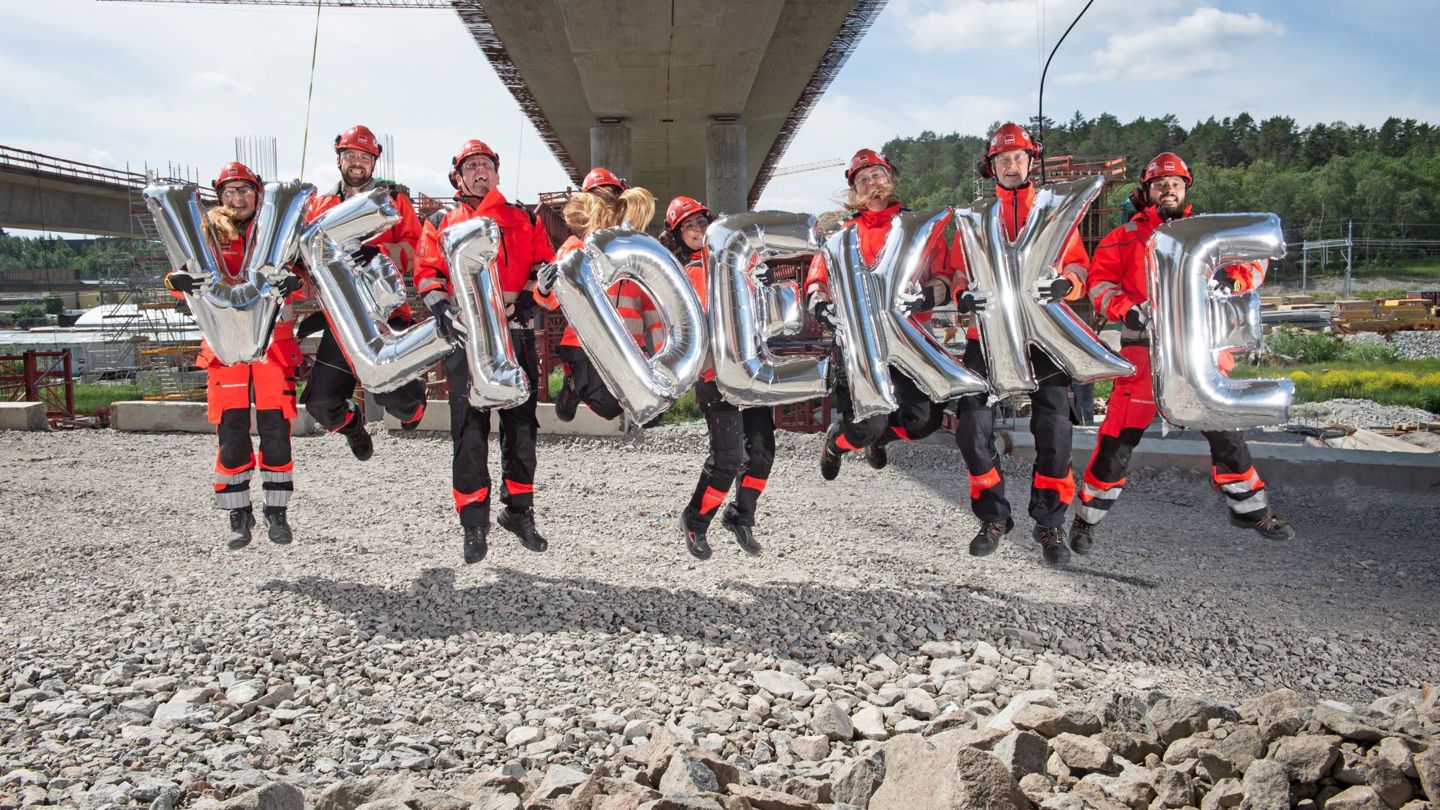 Under bro. Personer i varselkläder och hjälm hoppar glada med ballonger i famnen. Ballongerna bildar tillsammans ordet "Veidekke"