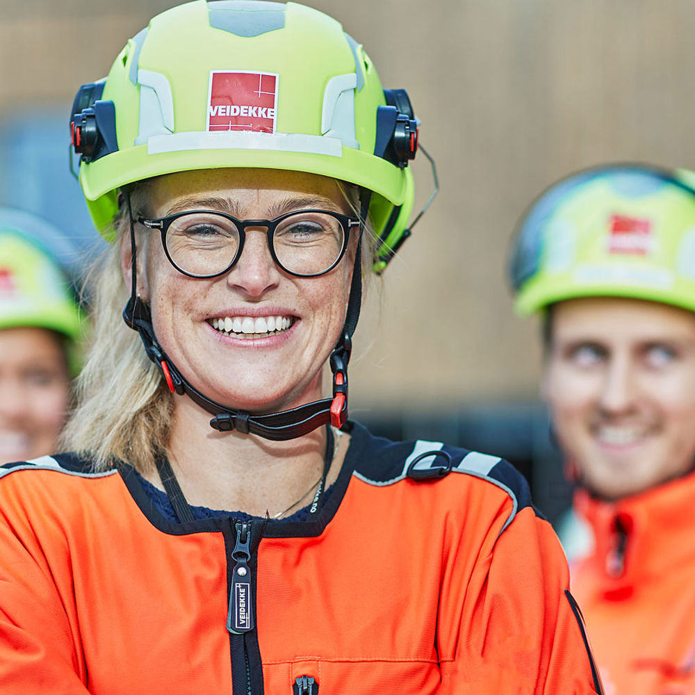 En tjej i varselkläder står leende med två kollegor i bakgrunden.