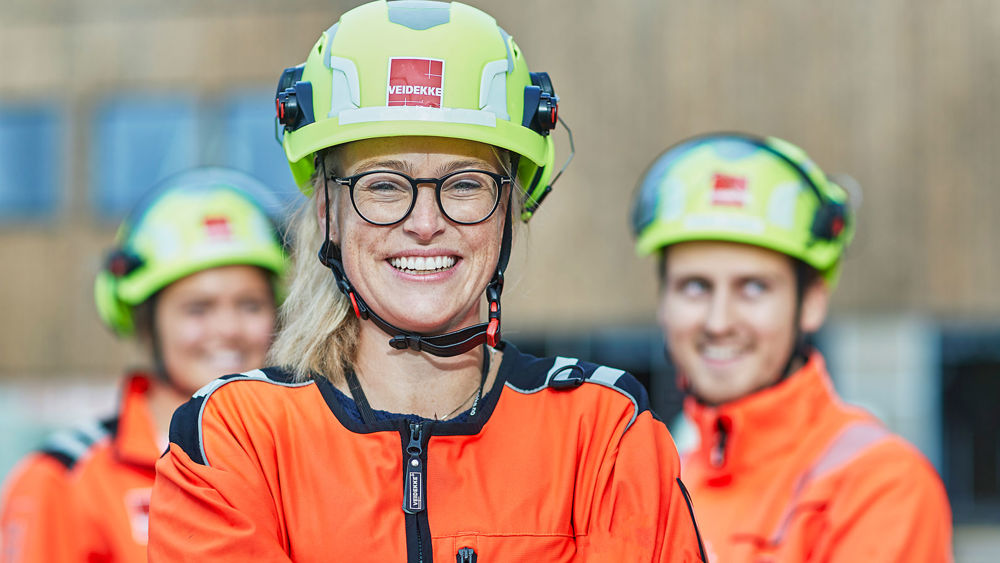 En tjej i varselkläder står leende med två kollegor i bakgrunden.