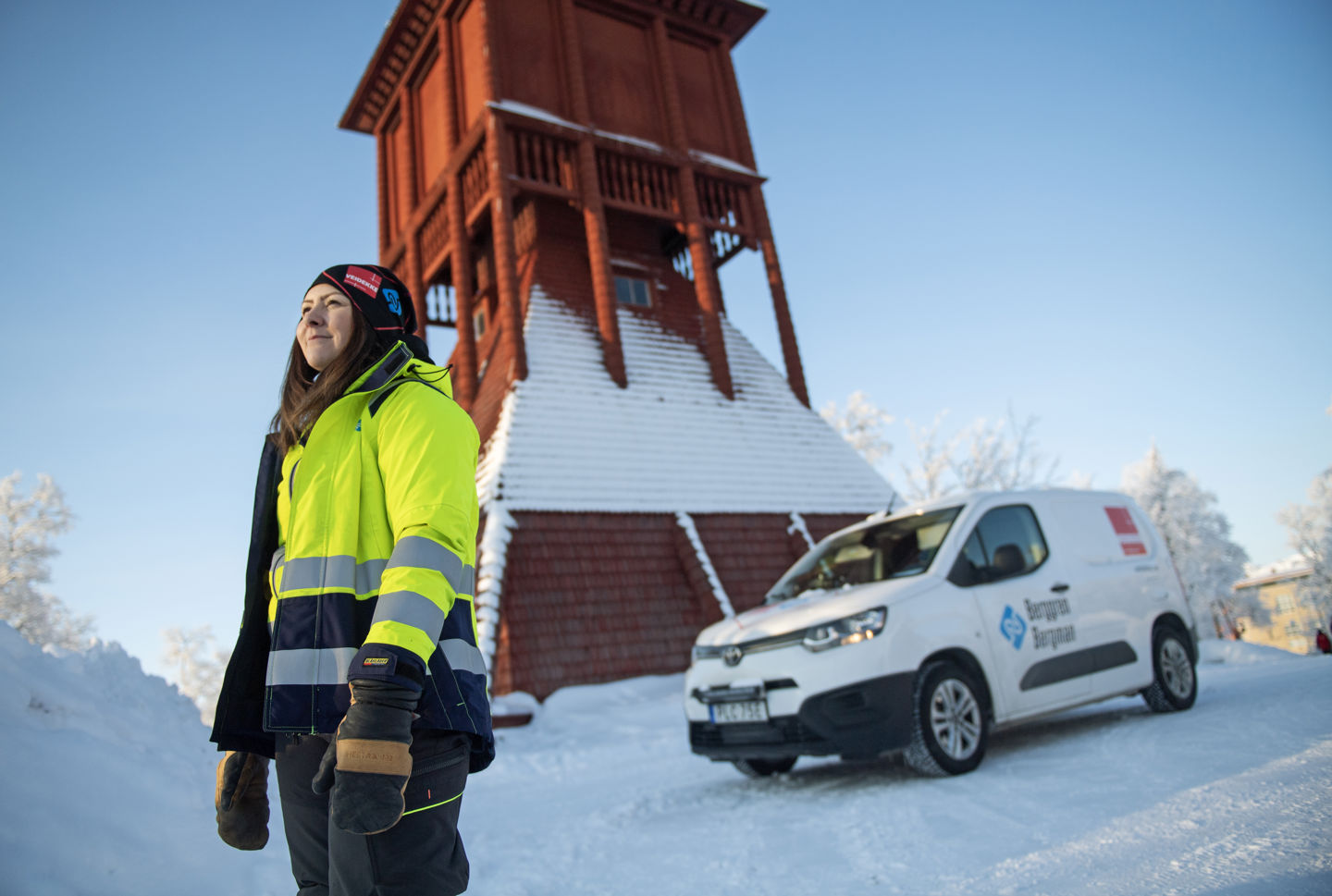 Rött kyrktorn i trä med snö på. Framför står Malin Bergman i varselkläder bredvid en vit skåpbil. 