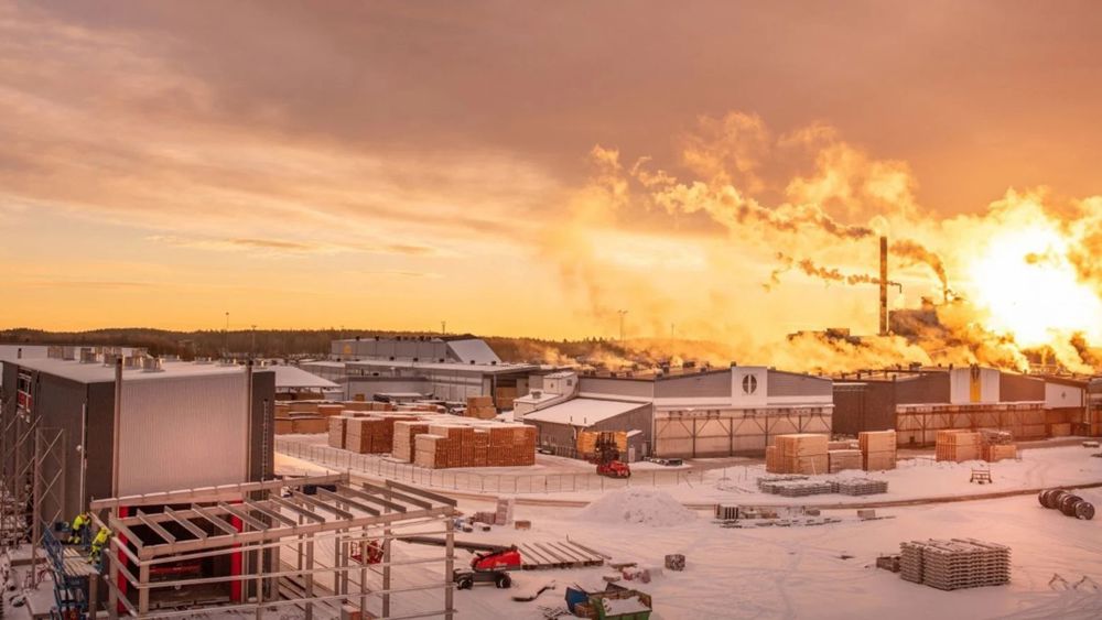 Vintervy över sågverket. Flera industribyygander med rykande skorstenar mot röd-gul himmel. Snö