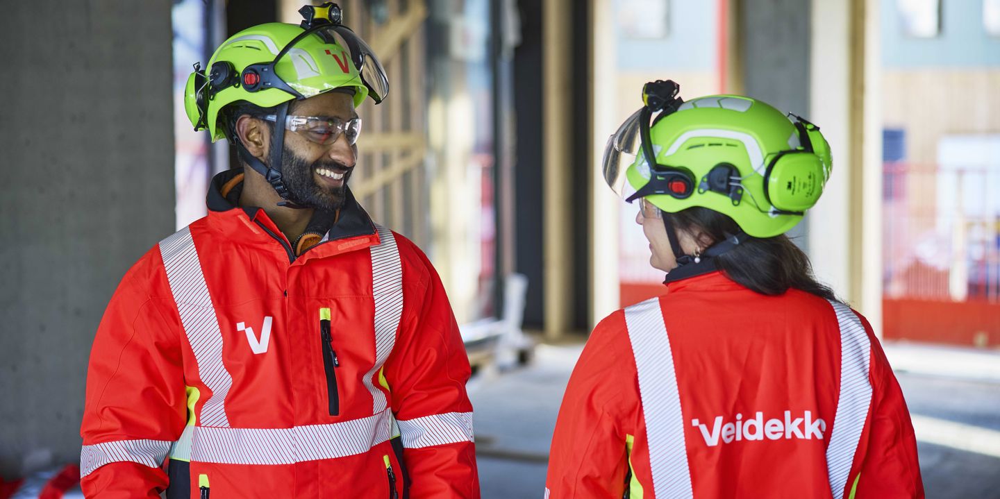 Två Veidekke-medarbetare i varselkläder ler mot varandra på en byggarbetsplats
