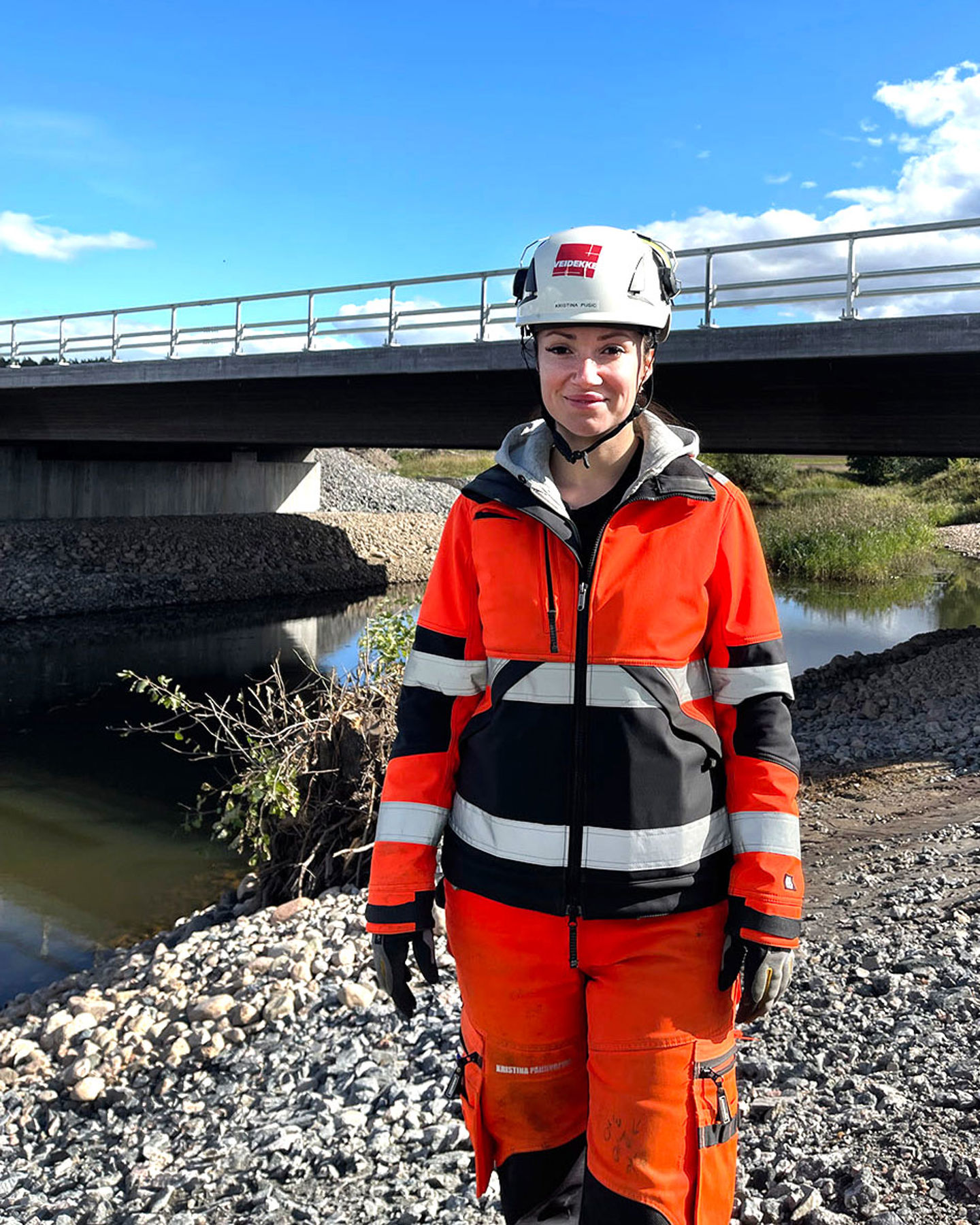 Tjej står leende framför en bro iklädd varselkläder och bygghjälm.