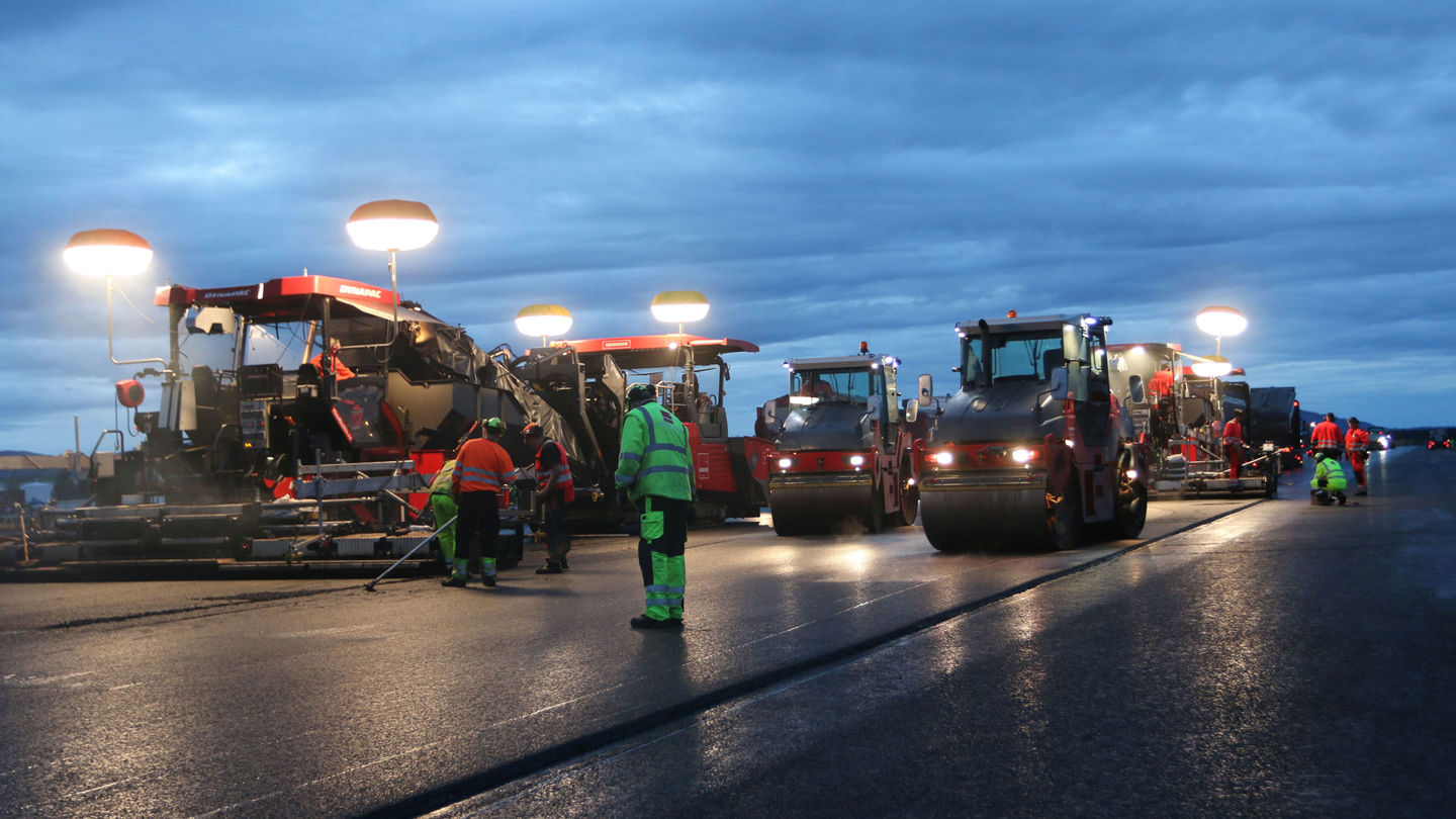 Flera stora maskiner arbetar med asfaltutläggning på en bred väg.