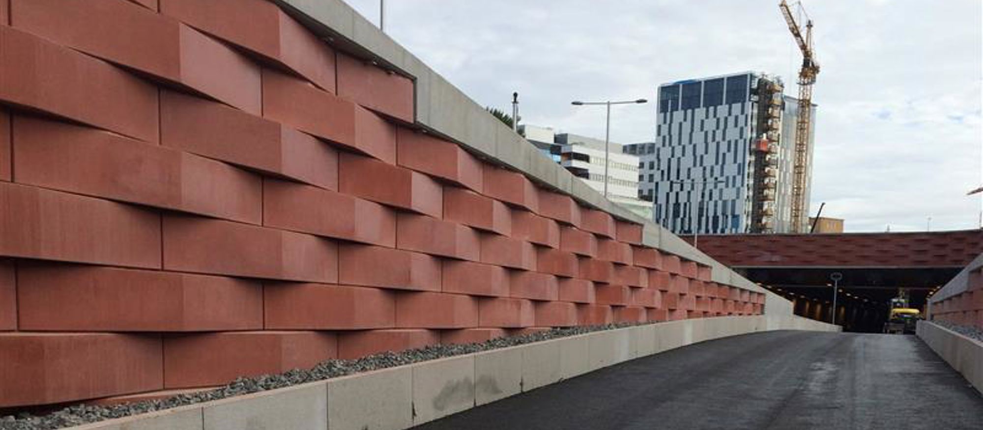Konstfärdigt utförd mur i terrakottafärgad betong vid motorväg. Höghus och byggkran i bakgrunden. 