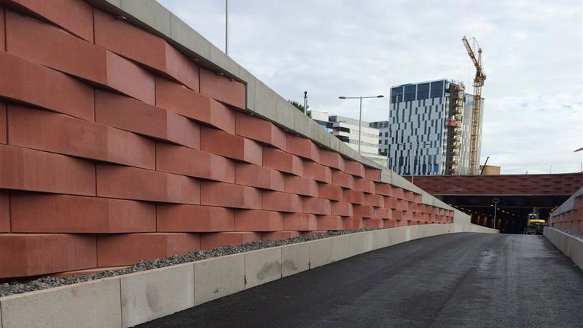 Konstfärdigt utförd mur i terrakottafärgad betong vid motorväg. Höghus och byggkran i bakgrunden. 
