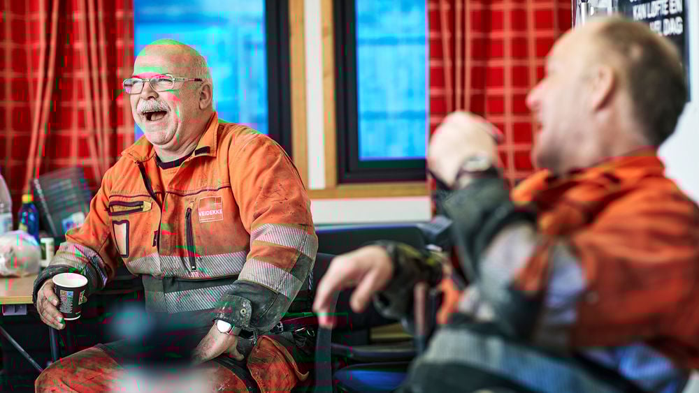 Två män på byggarbetsplats som skrattar och dricker kaffe.