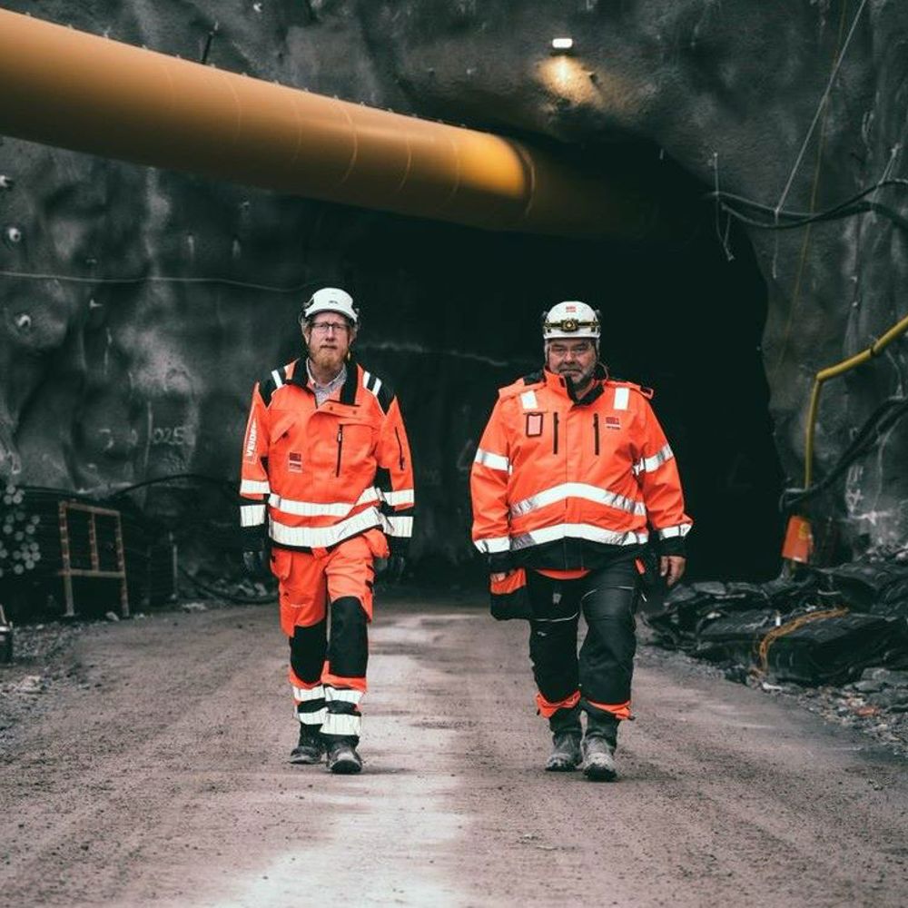 Bergtunnel med två män i varselkläder och tjockt gult rör ovanför