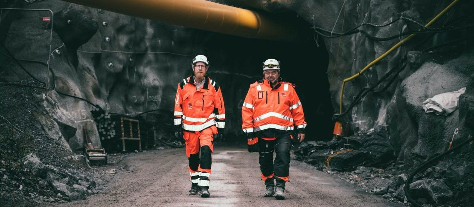 Bergtunnel med två män i varselkläder och tjockt gult rör ovanför