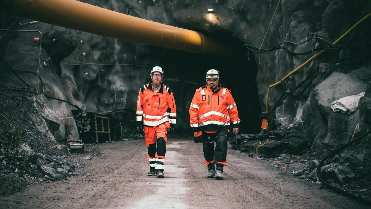 Bergtunnel med två män i varselkläder och tjockt gult rör ovanför