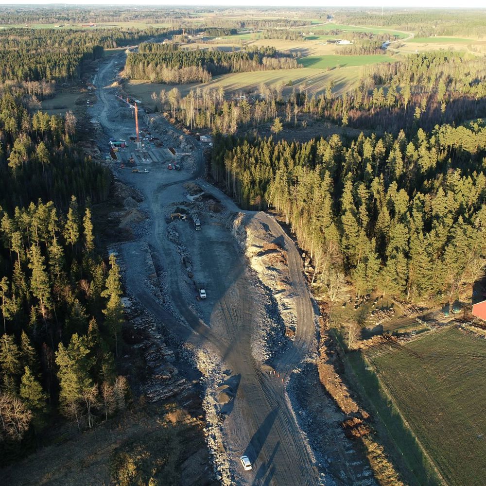 Drönarbild där vägsträckningen för E20 dras genom landskapet. Lastbil, en kran och början till brofundament. 