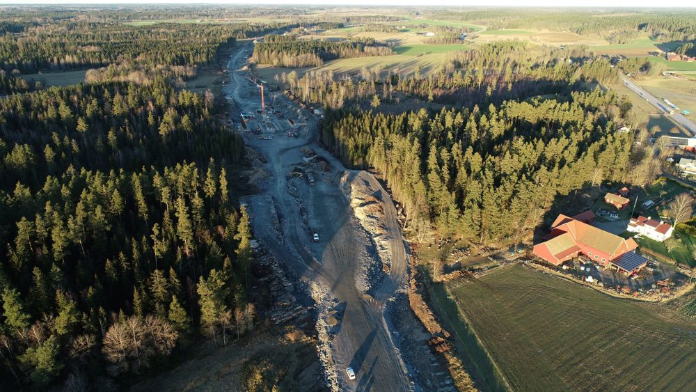 Drönarbild där vägsträckningen för E20 drogs genom landskapet. Lastbil, en kran och början till brofundament. 