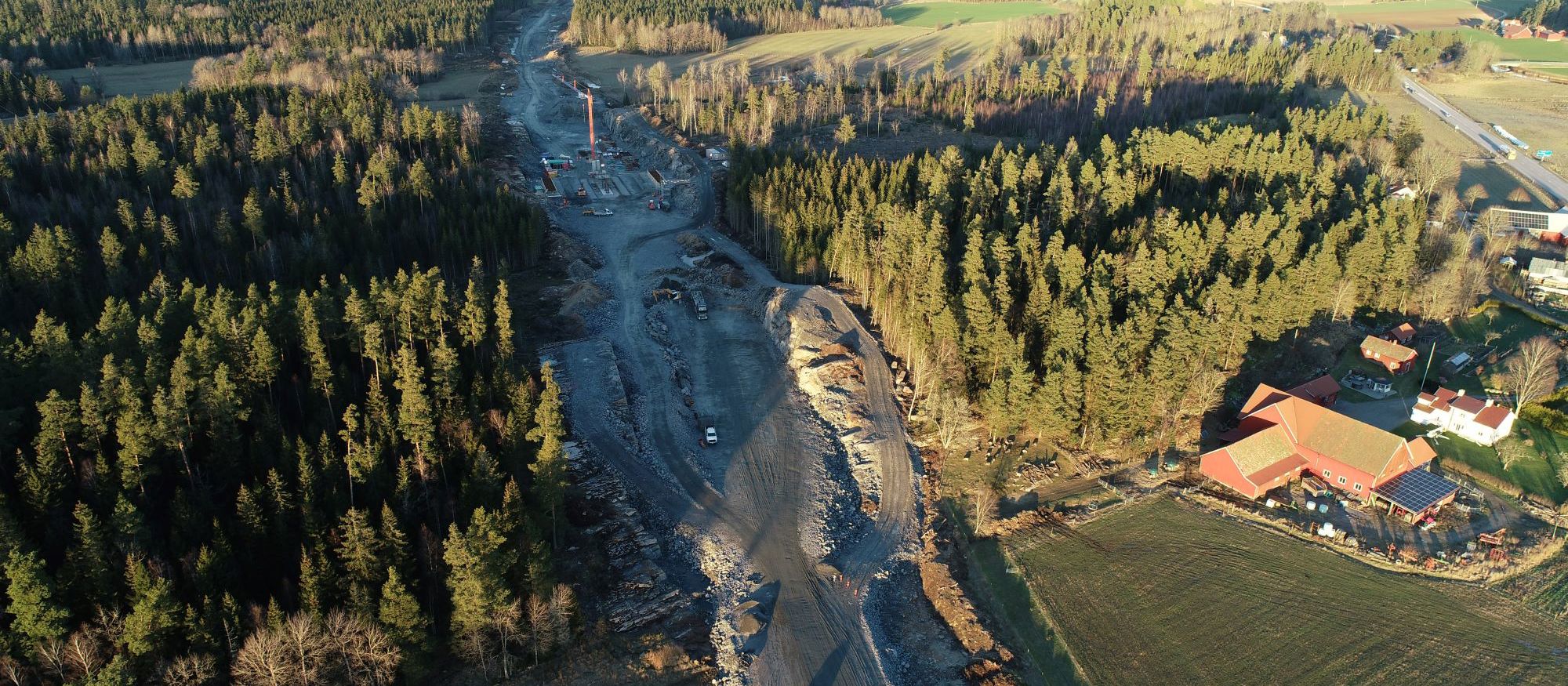 Drönarbild där vägsträckningen för E20 drogs genom landskapet. Lastbil, en kran och början till brofundament. 