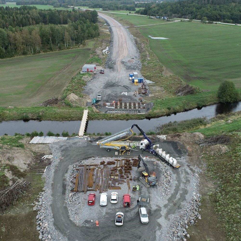 Pågående väggbygge där marken jämnats. Balkar och betongfundament förbereds för att bygga bro över en å i förgrunden.