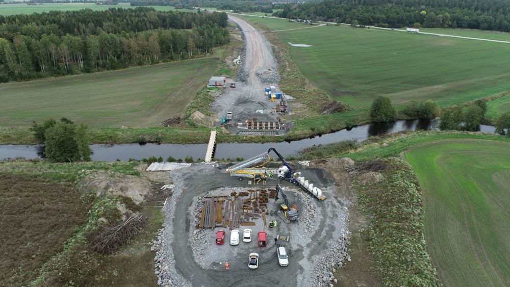 Pågående väggbygge där marken jämnats. Balkar och betongfundament förbereds för att bygga bro över en å i förgrunden.
