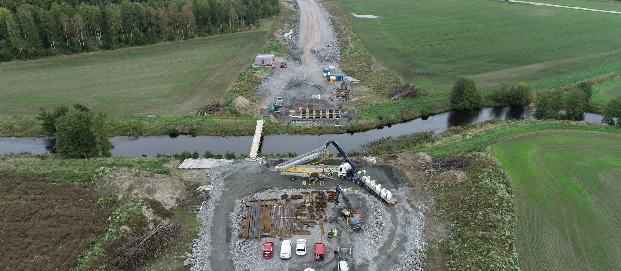 Pågående väggbygge där marken jämnats. Balkar och betongfundament förbereds för att bygga bro över en å i förgrunden.