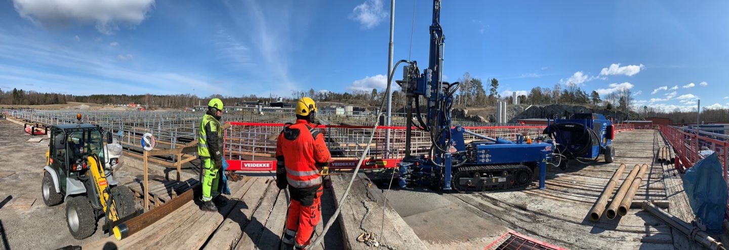 Arbete pågår vid Himmerfjärdsverket. Anläggningsarbetare och maskiner i förgrunden i gula och röda varselkläder. Bassänger i bakgrunden. Blå himmel.