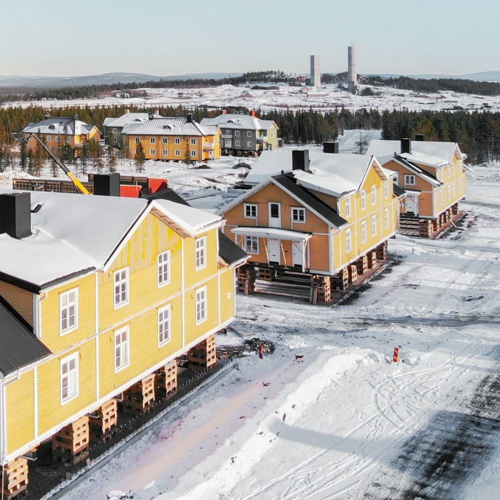 Vi flyttar “Gula raden” i Kiruna. Tre stora gula trähus har lyfts upp på pallar ovanför marken i snöig omgivning. 